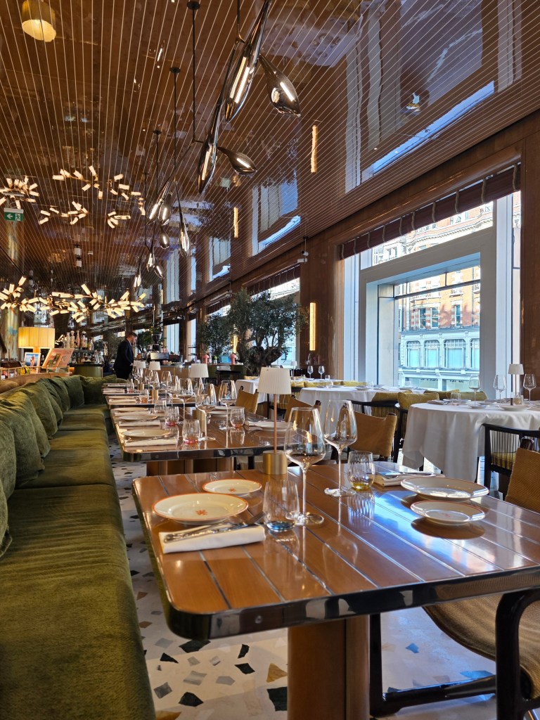 Interior of a modern restaurant featuring a long green sofa and elegantly set tables with glassware, surrounded by ambient lighting and large windows.