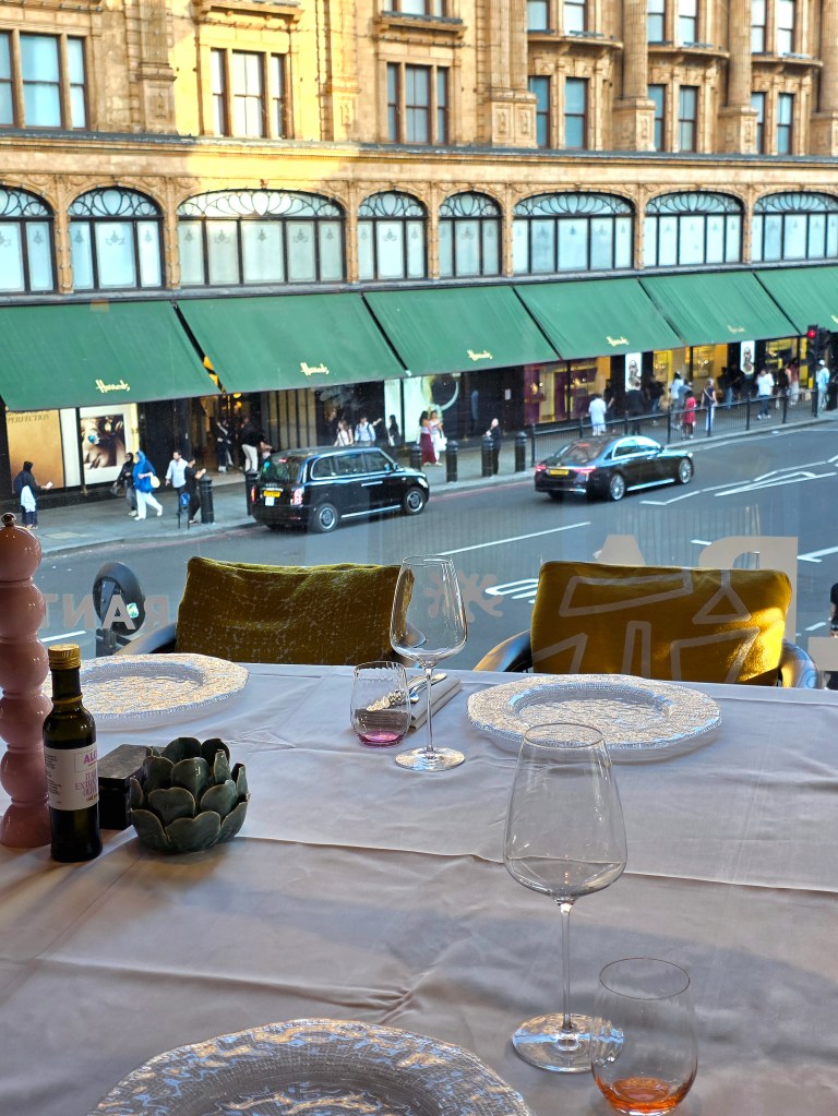 A table set for dining with elegant plates, glasses, and olive oil, overlooking a bustling street with shops and people outside.