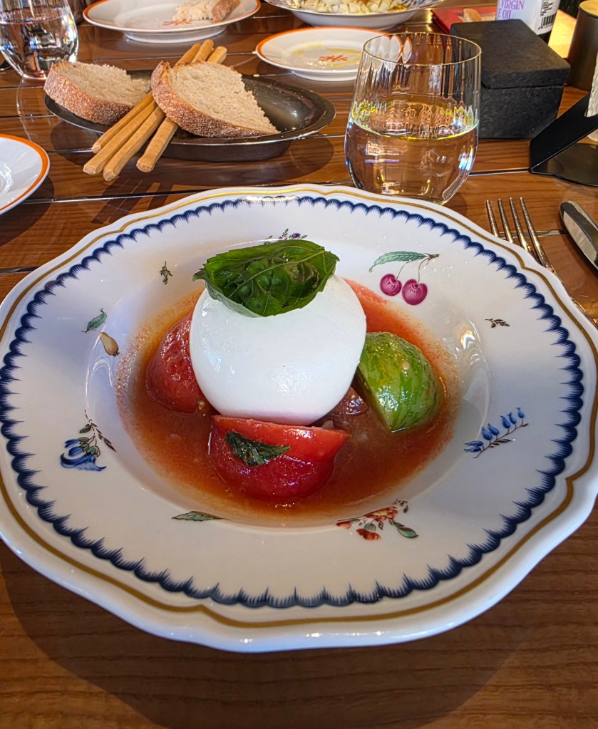 A decorated plate featuring a large ball of burrata cheese, surrounded by slices of red and green tomatoes, in a light tomato broth with a basil leaf garnish.