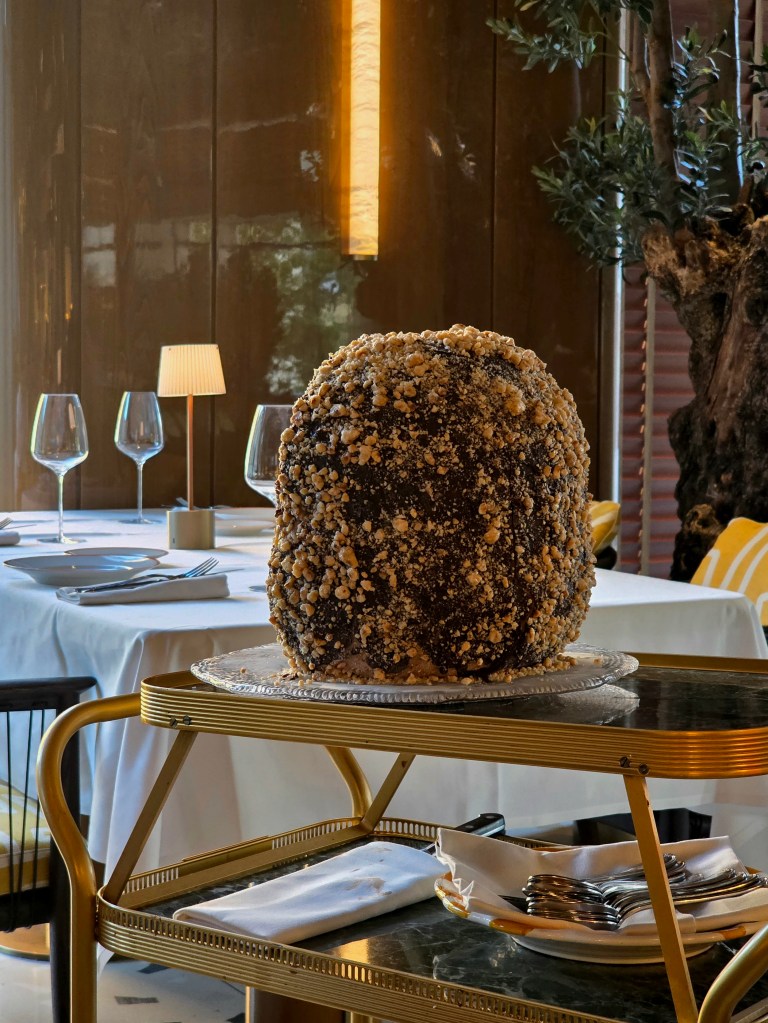 A large, elaborately decorated dessert with a dark chocolate exterior and a crunchy topping of chopped nuts, displayed on a serving trolley in an elegant restaurant setting.