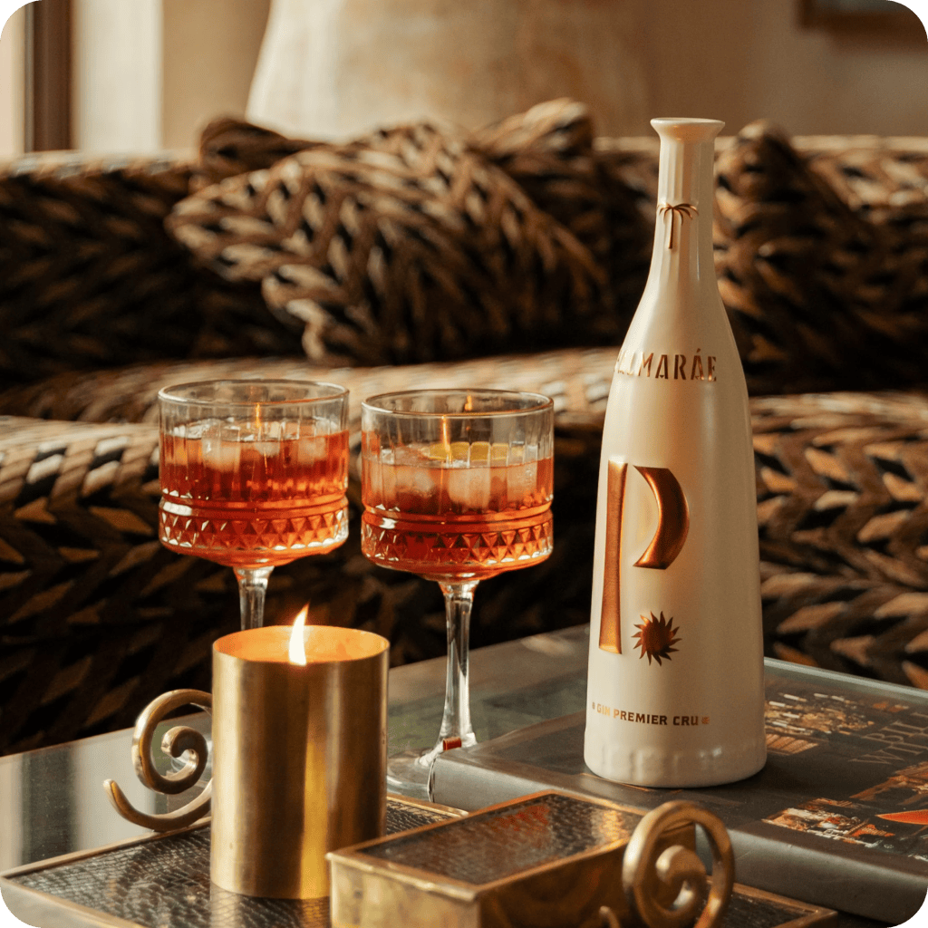 A stylish table setting featuring two elegant glasses of red cocktails, a decorative candle in a gold holder, and a bottle of gin on a wooden surface, with a patterned sofa in the background.