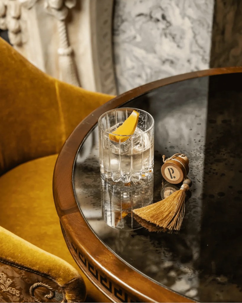 A glass of cocktail with ice and a lemon wedge sits on a glossy black table, alongside a decorative tassel and a wooden charm.