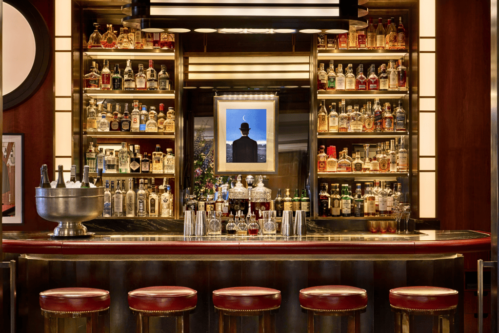 A stylish bar interior featuring a polished countertop, multiple shelves filled with various bottles of spirits, an ornate ice bucket, and four red cushioned stools. A framed artwork depicting a figure in a hat is visible on the wall, adding to the sophisticated atmosphere.
