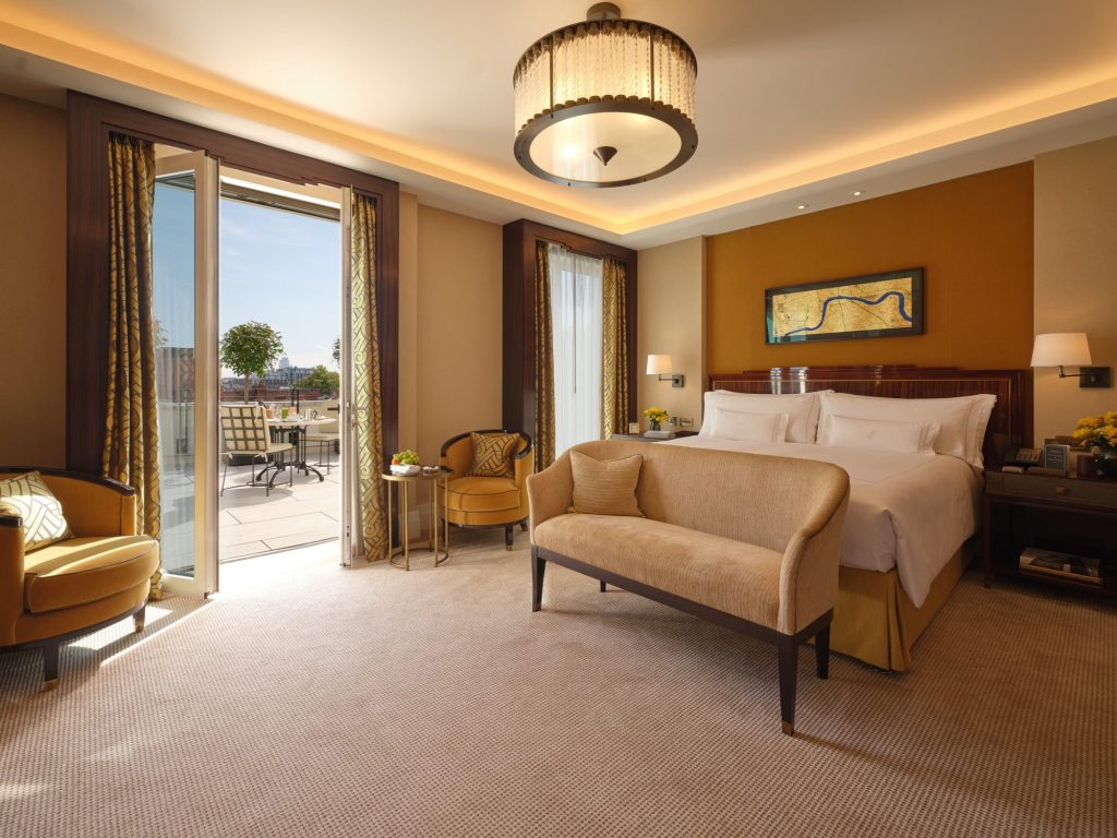 A luxurious hotel room featuring a comfortable bed, stylish seating area with gold armchairs, and a private balcony with outdoor seating overlooking a scenic view, well-lit with natural light and elegant decor.