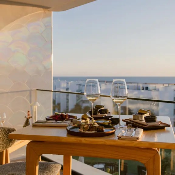 A dining table set on a balcony overlooking the sea at sunset, featuring two glasses of wine, assorted dishes, and elegant tableware.