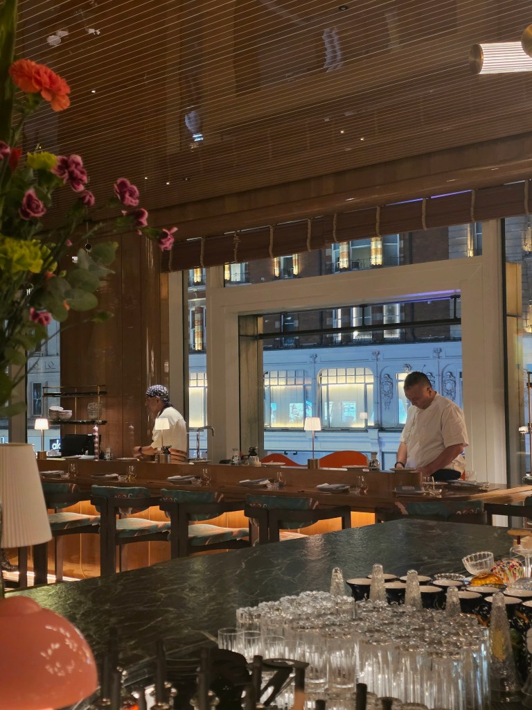 View of a restaurant interior with two chefs preparing at a counter, surrounded by elegant decor and dining tables, with city lights visible through large windows.