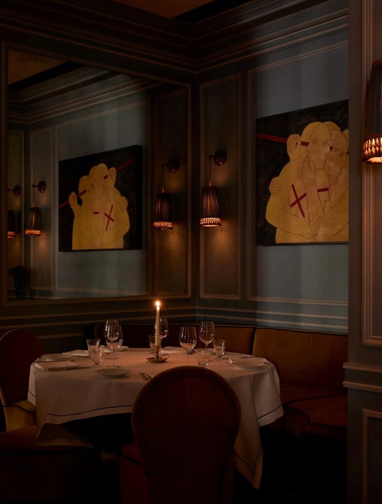 Intimate restaurant setting featuring a round table with a white tablecloth and candlelit centrepiece, surrounded by plush seating. The dimly lit room has wall art and decorative lighting.