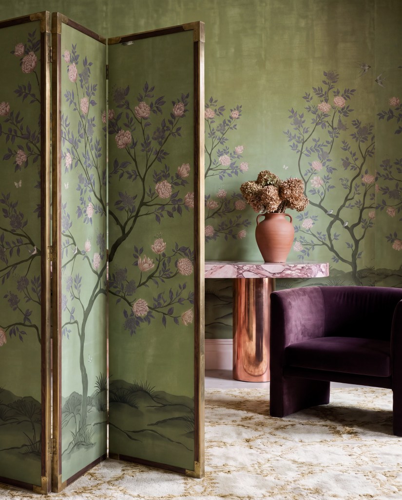 A stylish interior scene featuring a decorative folding screen with floral designs, a modern velvet chair, a pink marble side table, and a terracotta vase with dried flowers against a green wallpaper backdrop.