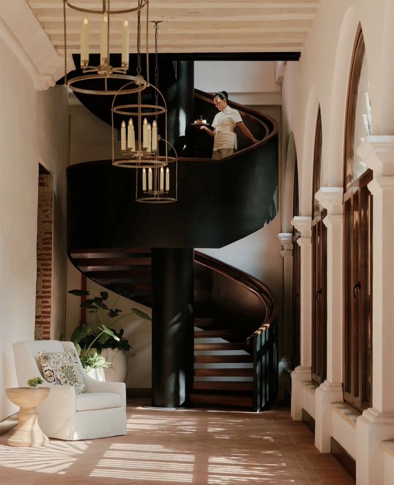 A modern and elegant spiral staircase in a spacious interior, featuring a person carrying a tray. The scene includes stylish hanging light fixtures, a comfortable armchair with a patterned cushion, and potted plants, all bathed in natural light.