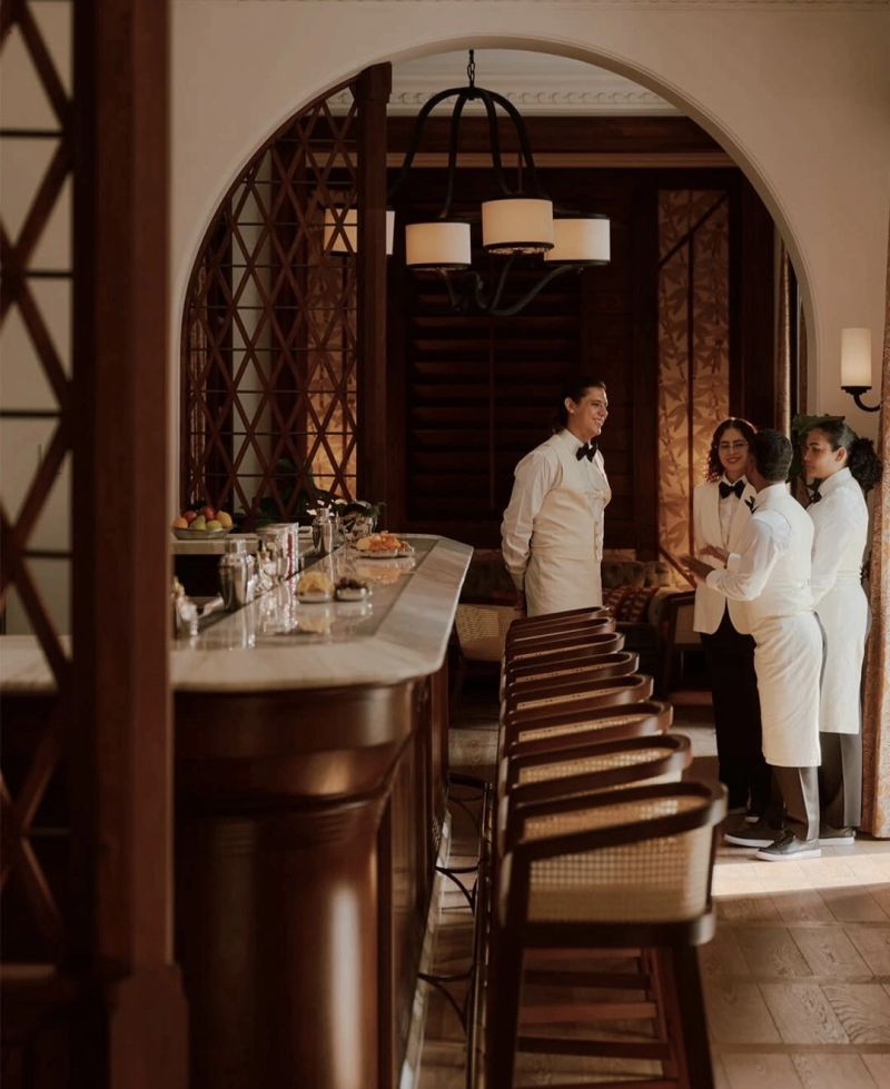 A stylish bar area featuring a marble countertop and wooden seating, with four staff members in formal attire engaged in conversation.