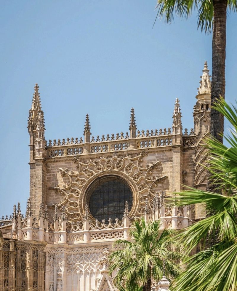 Close-up view of the ornate architecture and decorative elements of a historic building, featuring a large circular window and pointed spires, with palm trees in the foreground and a clear blue sky.