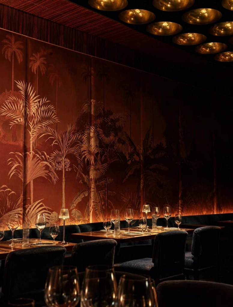 Interior of a stylish restaurant featuring a tropical wallpaper design, elegantly set tables with glassware, and atmospheric lighting.