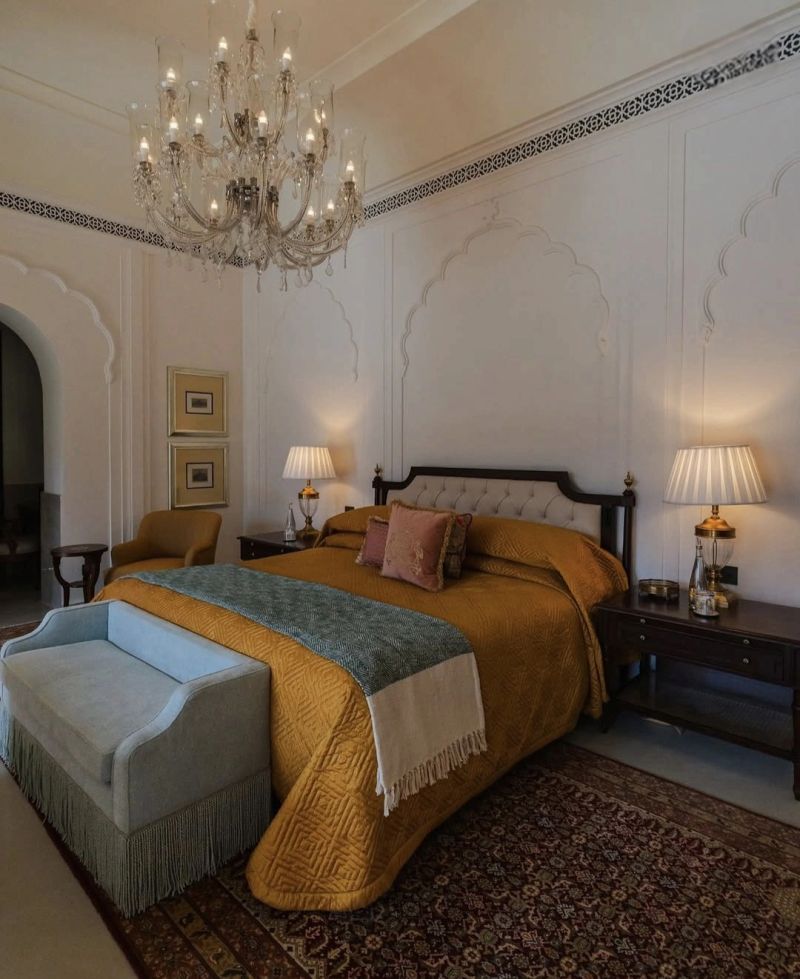 Elegant bedroom featuring a large bed with a mustard yellow coverlet, decorative pillows, and a light blue bench at the foot. A crystal chandelier hangs from the ceiling, complemented by two bedside lamps on wooden tables. The walls are adorned with intricate patterns, and an ornate rug adds warmth to the space.