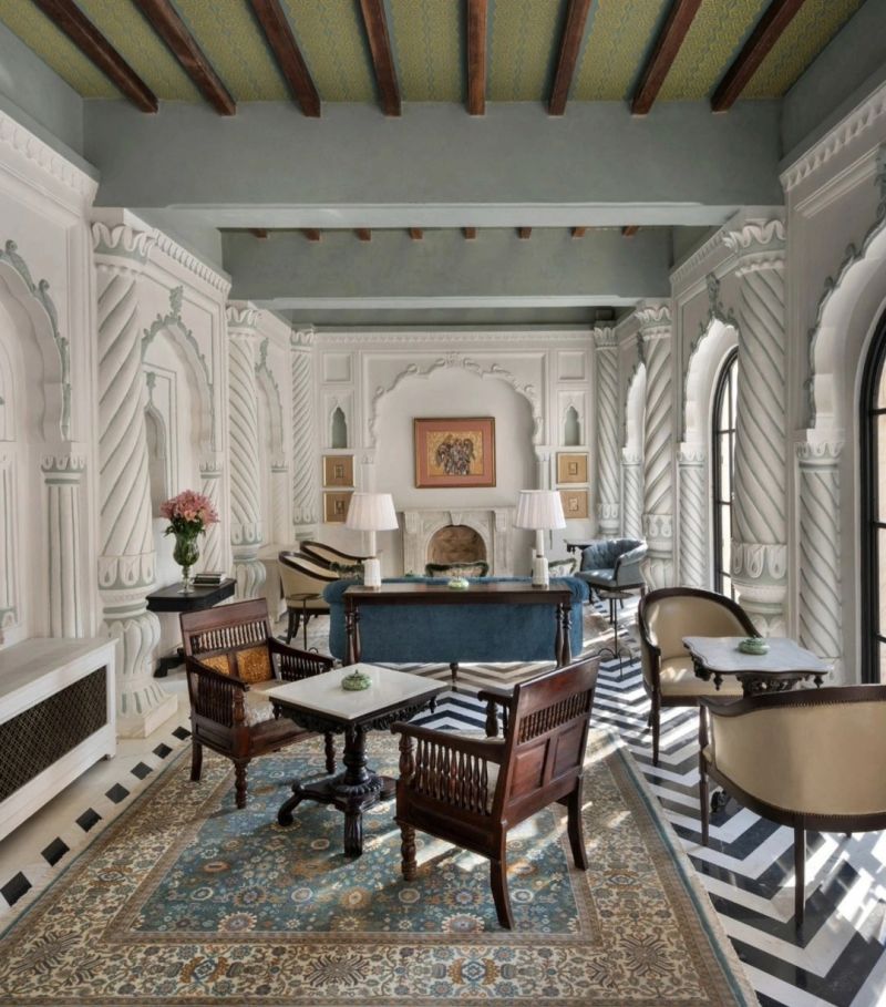 A stylish and elegant living room featuring intricate white moulded walls, wooden beams on the ceiling, vintage furniture, and a decorative rug. The room includes two armchairs, wooden table, and a fireplace, creating a warm and inviting atmosphere.