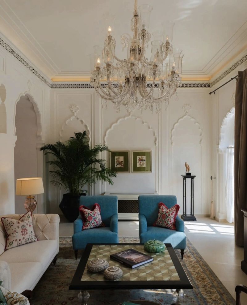 A stylish living room featuring a crystal chandelier, two blue armchairs with patterned cushions, a light sofa adorned with floral pillows, and a decorative coffee table. The walls are adorned with elegant arches and framed artwork, with a lush plant adding greenery to the space.