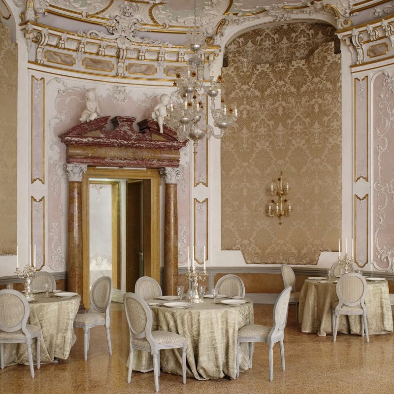 Elegant dining room featuring ornate ceilings, decorative wall patterns, and tables set with tablecloths and fine dinnerware.