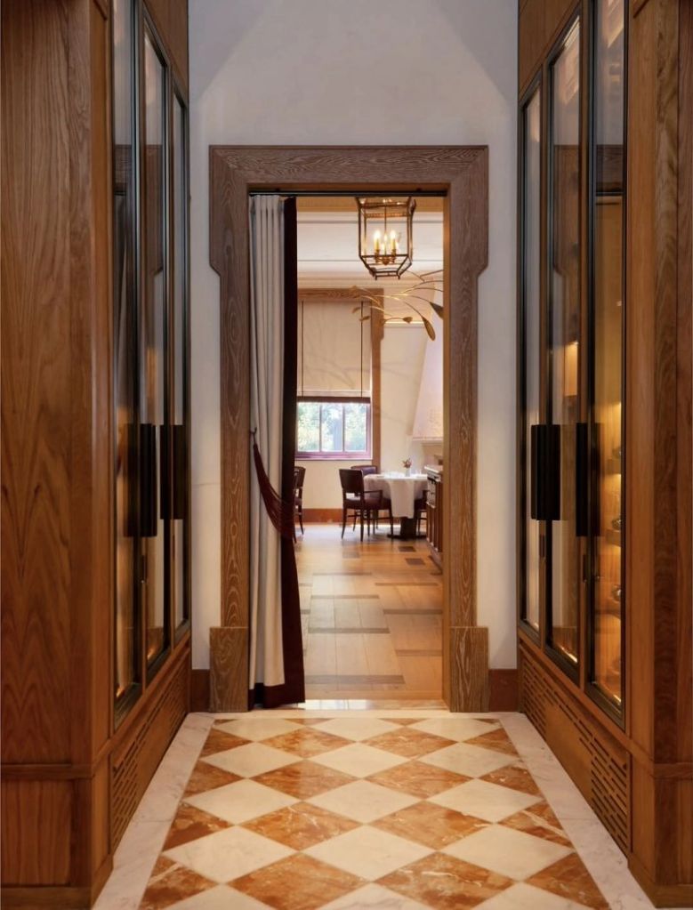 A hallway with wooden paneling, featuring glass-fronted cabinets on either side and a patterned marble floor. At the end of the hallway, a table with chairs is visible in a well-lit dining area.