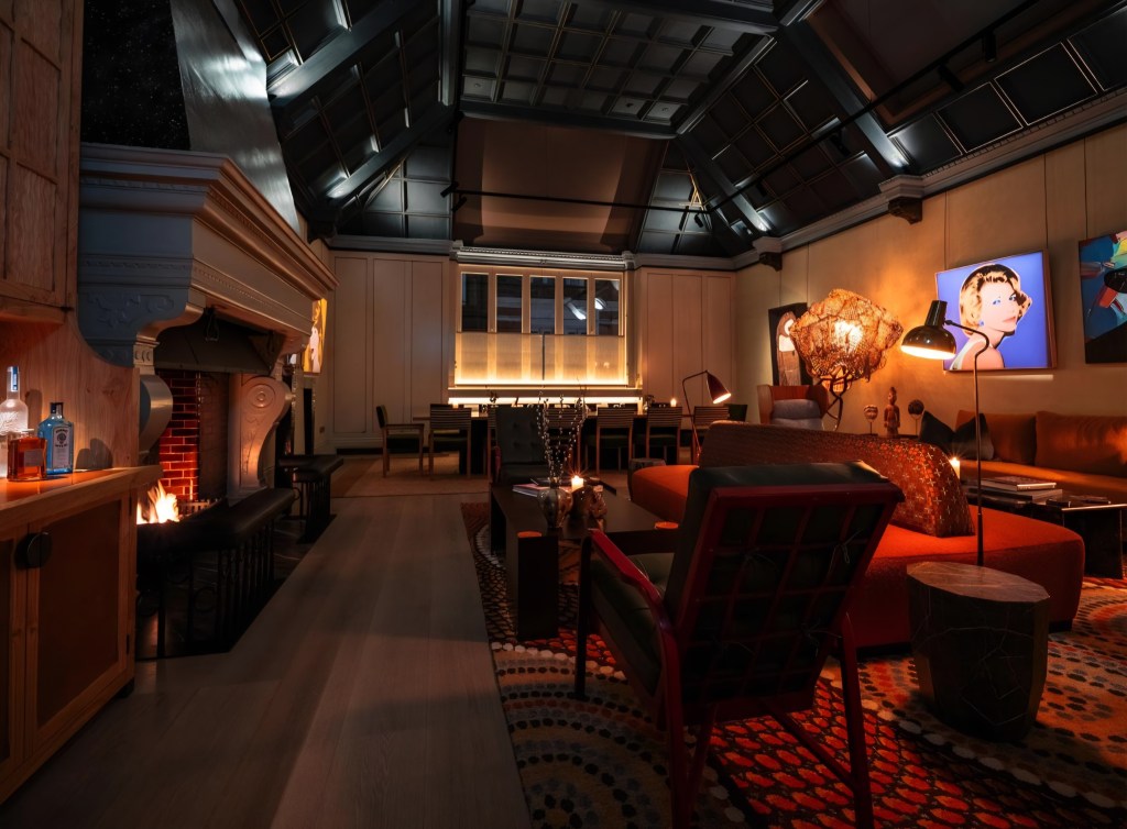 A stylish, modern living room featuring a high ceiling, a warm fireplace, and contemporary art on the walls. The room is furnished with plush seating, wooden accents, and ambient lighting, creating a cosy yet sophisticated atmosphere.